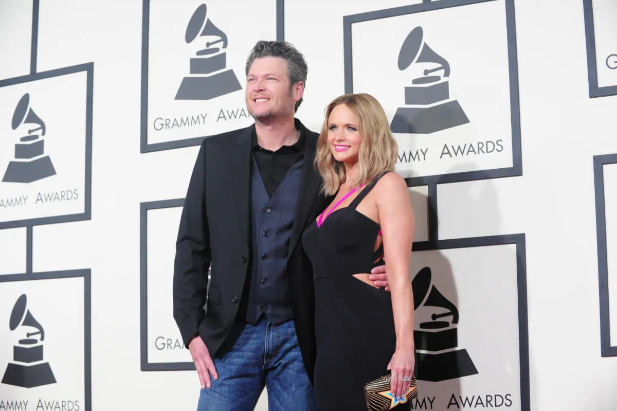 Blake Shelton stands with his arm around Miranda Lambert. He wears jeans and a blazer and she wears a black dress.