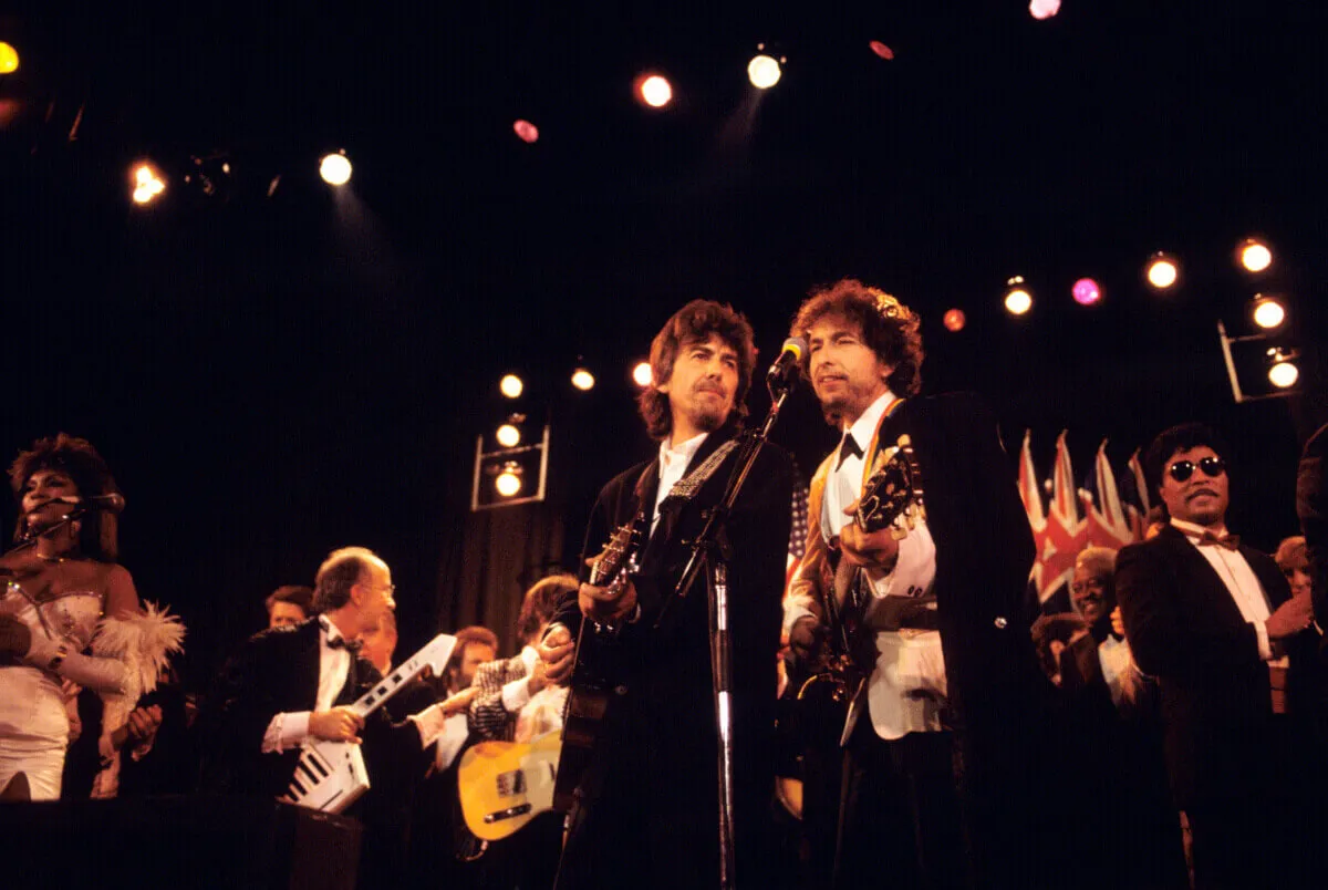 George Harrison and Bob Dylan stand in front of a microphone on a stage.