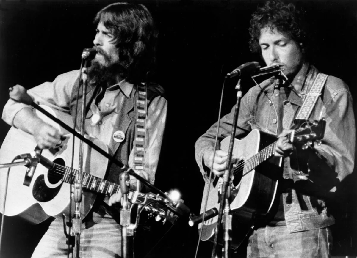 A black and white picture of George Harrison and Bob Dylan standing in front of microphones with acoustic guitars. Dylan has a harmonica.