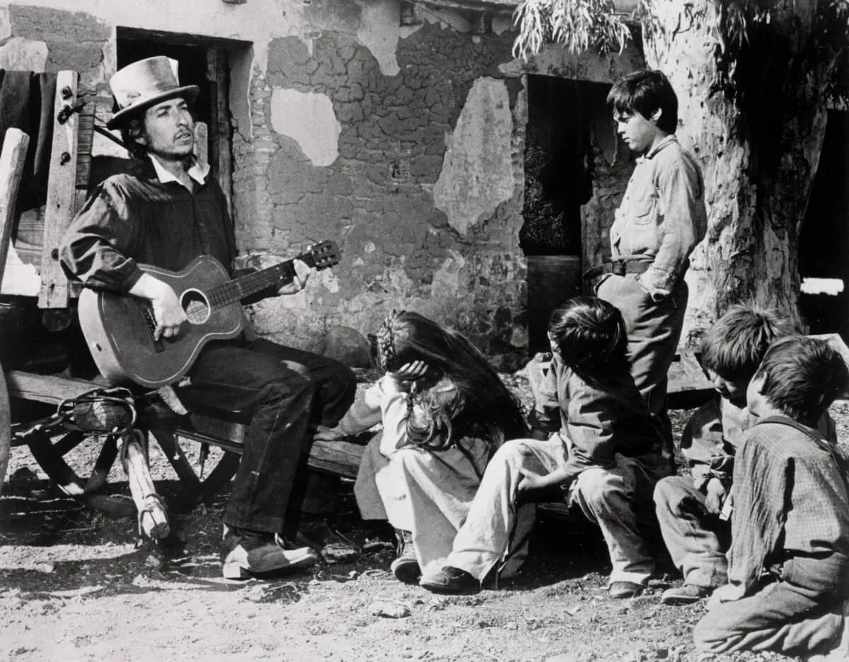 A black and white picture of Bob Dylan sitting and playing guitar for a group of children.