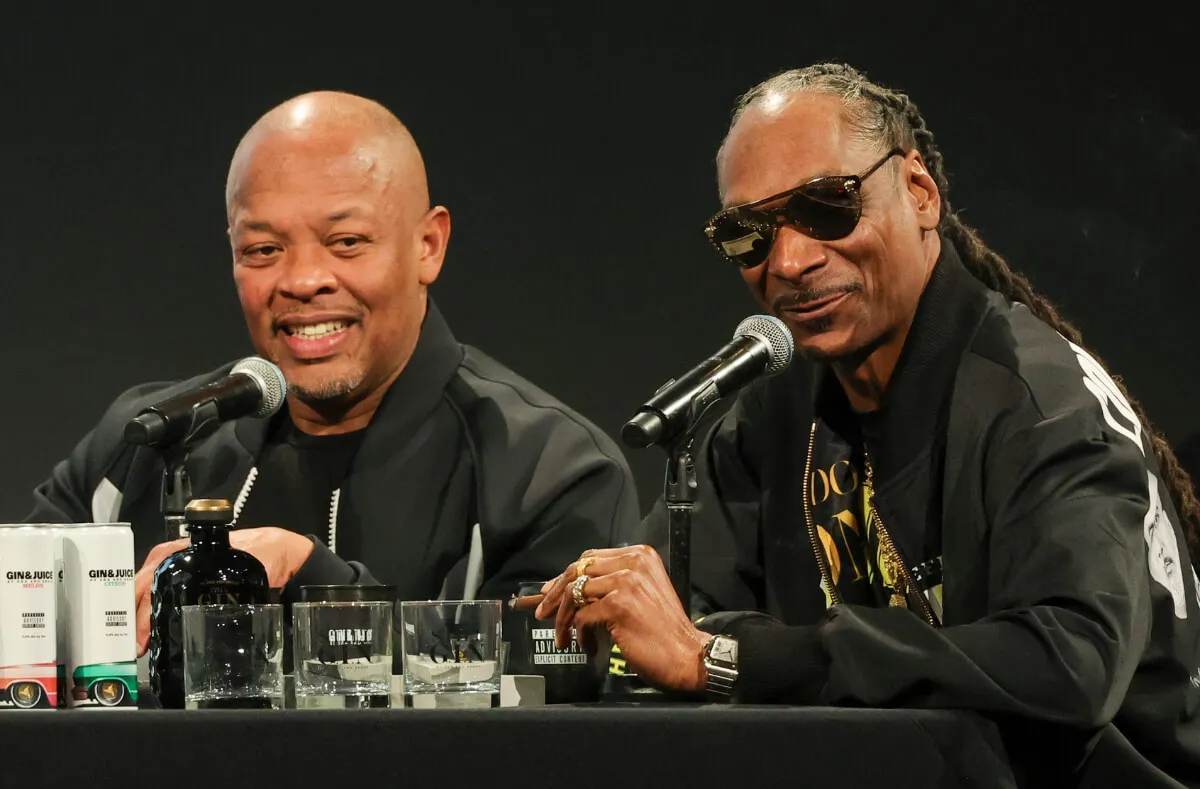 Dr. Dre and Snoop Dogg sit together with microphones in front of them. They both wear black, and Snoop has sunglasses on.