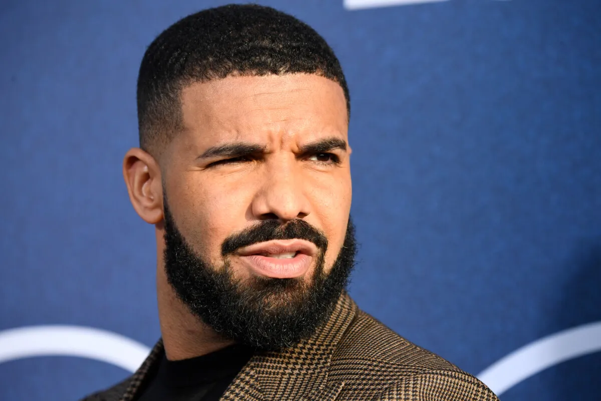 Drake wears a brown jacket and stands in front of a blue background.