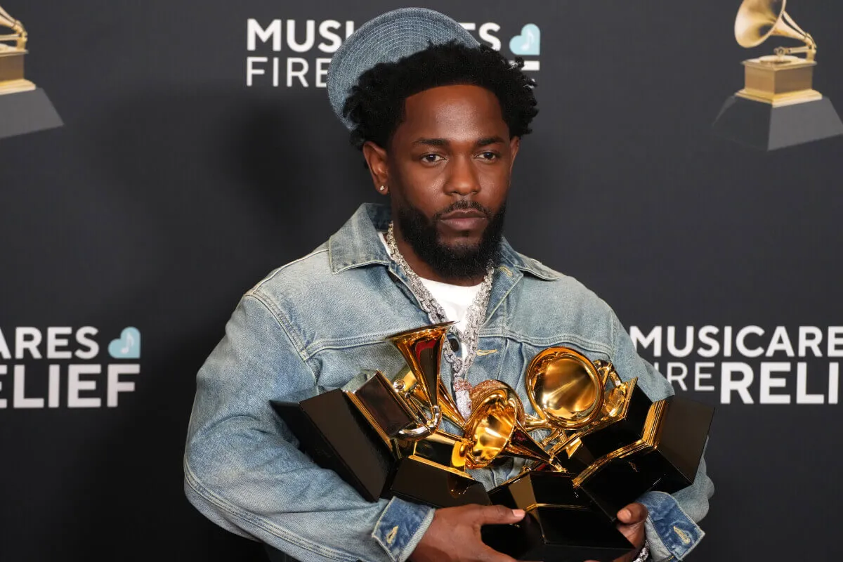 Kendrick Lamar carries a number of Grammys while wearing a denim jacket and hat.