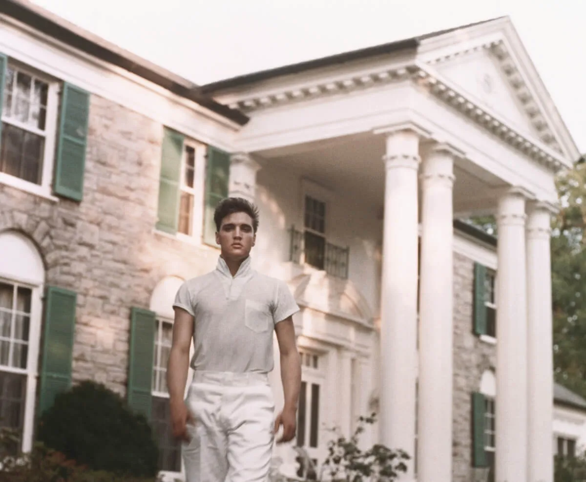 Elvis wears a white shirt and white pants. He stands in front of Graceland.