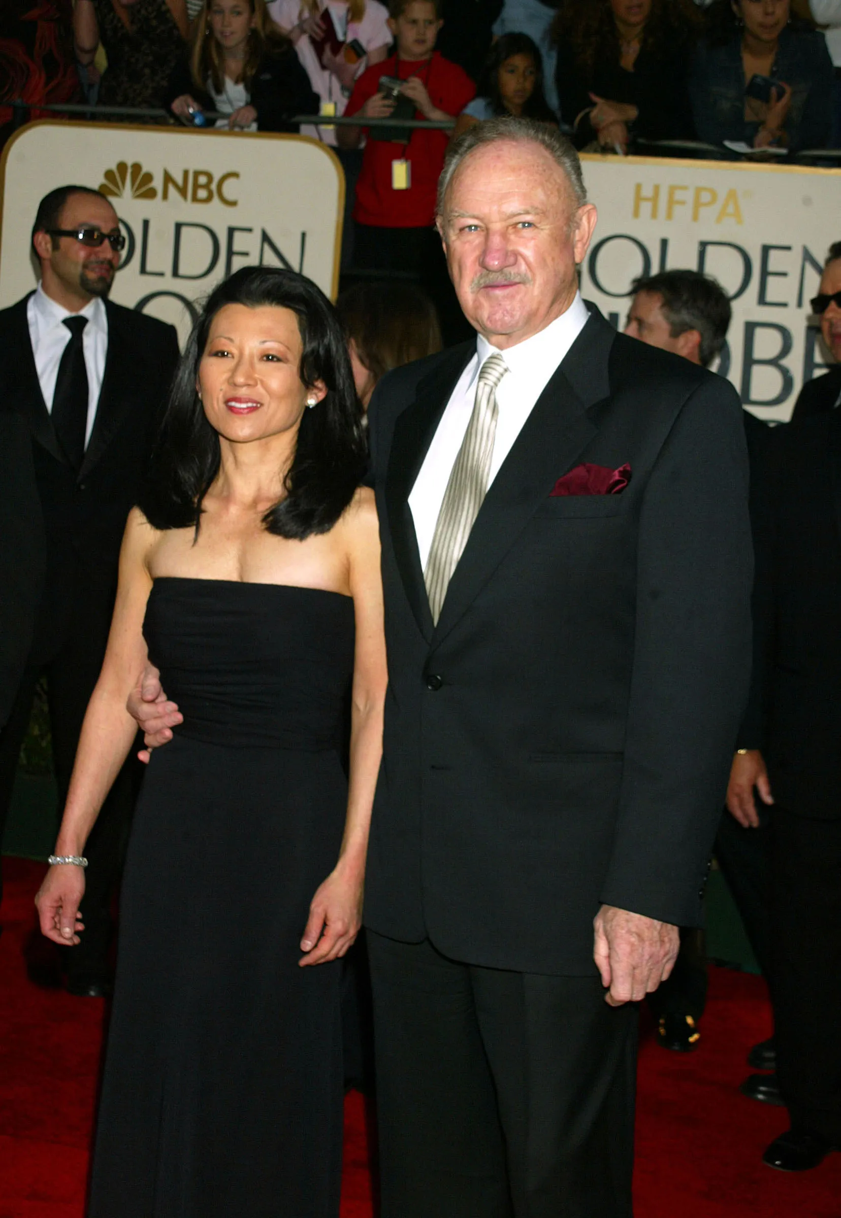 Gene Hackman and wife Betsy Arakawa at the 60th Annual Golden Globe Awards. Hackman is wearing a black suit and has his arm around his wife, who is wearing a black sleeveless dress.