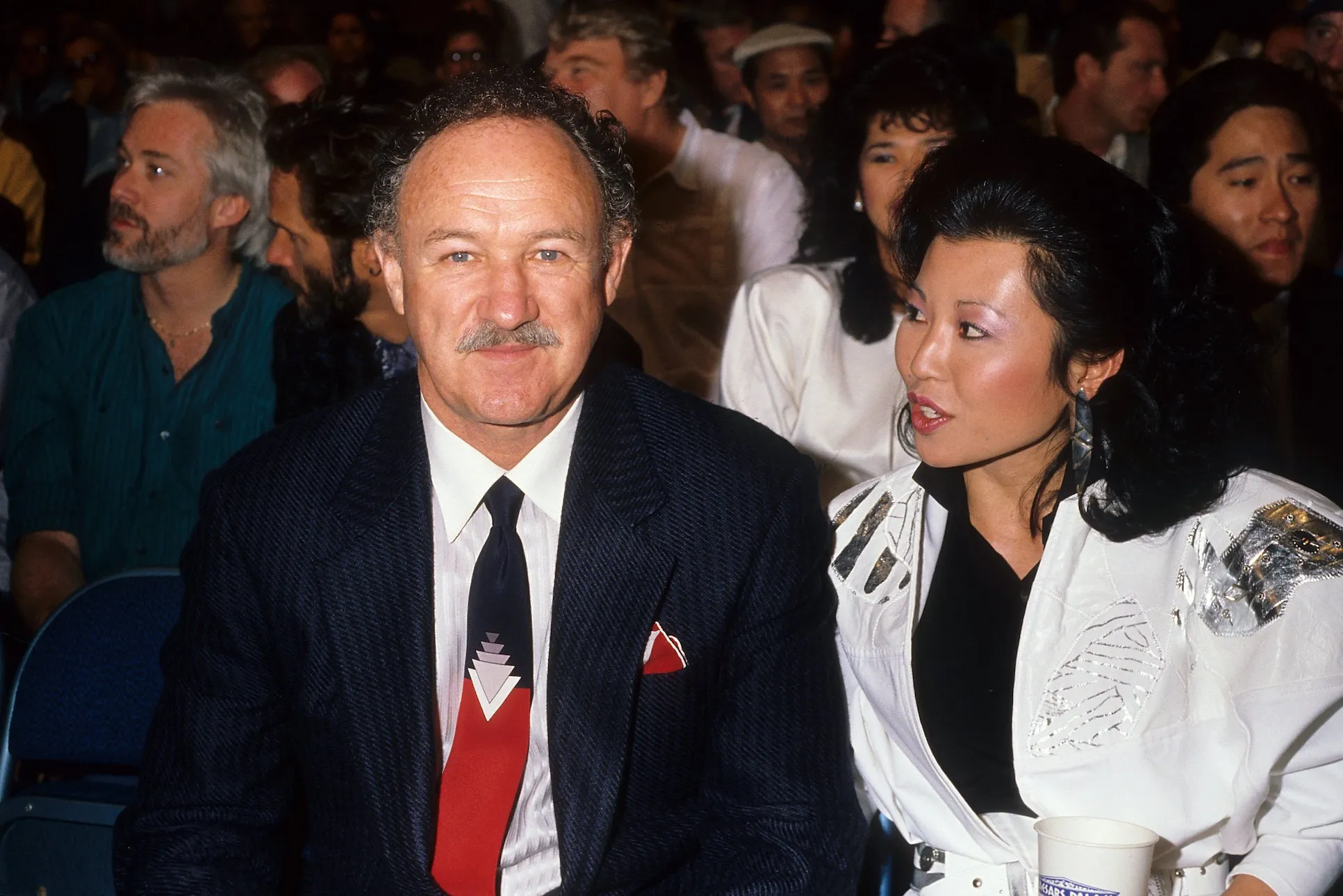 Gene Hackman and Betsy Arakawa sitting next to each other in 1986. Hackman is looking forward while Arakawa is looking at him.