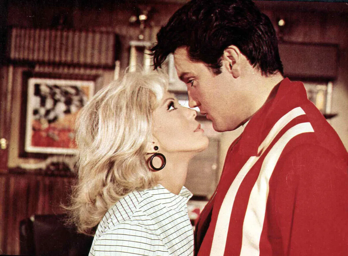 Nancy Sinatra and Elvis Presley stand with their faces close together. She wears a white shirt with black stripes and he wears a red jacket.