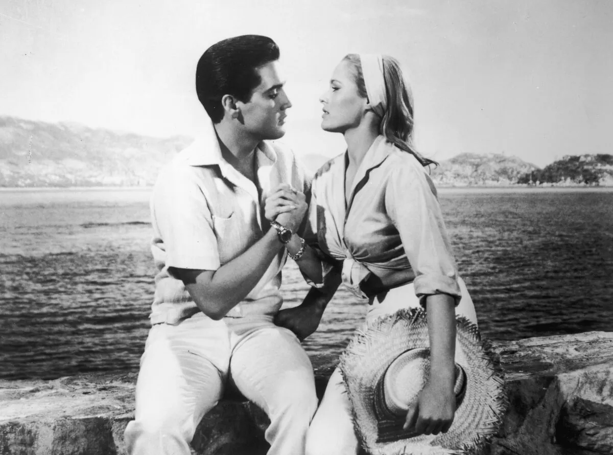 A black and white picture of Elvis Presley clasping Ursula Andress' hand. They sit in front of water.