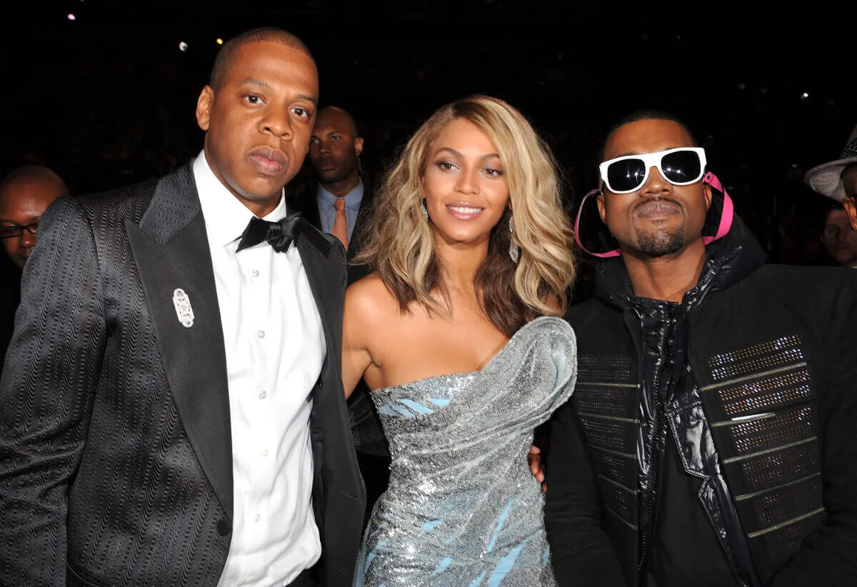 Beyonce wears a silver dress and stands between Jay-Z, who wears a tuxedo, and Kanye West, who wears a black jacket and white sunglasses.