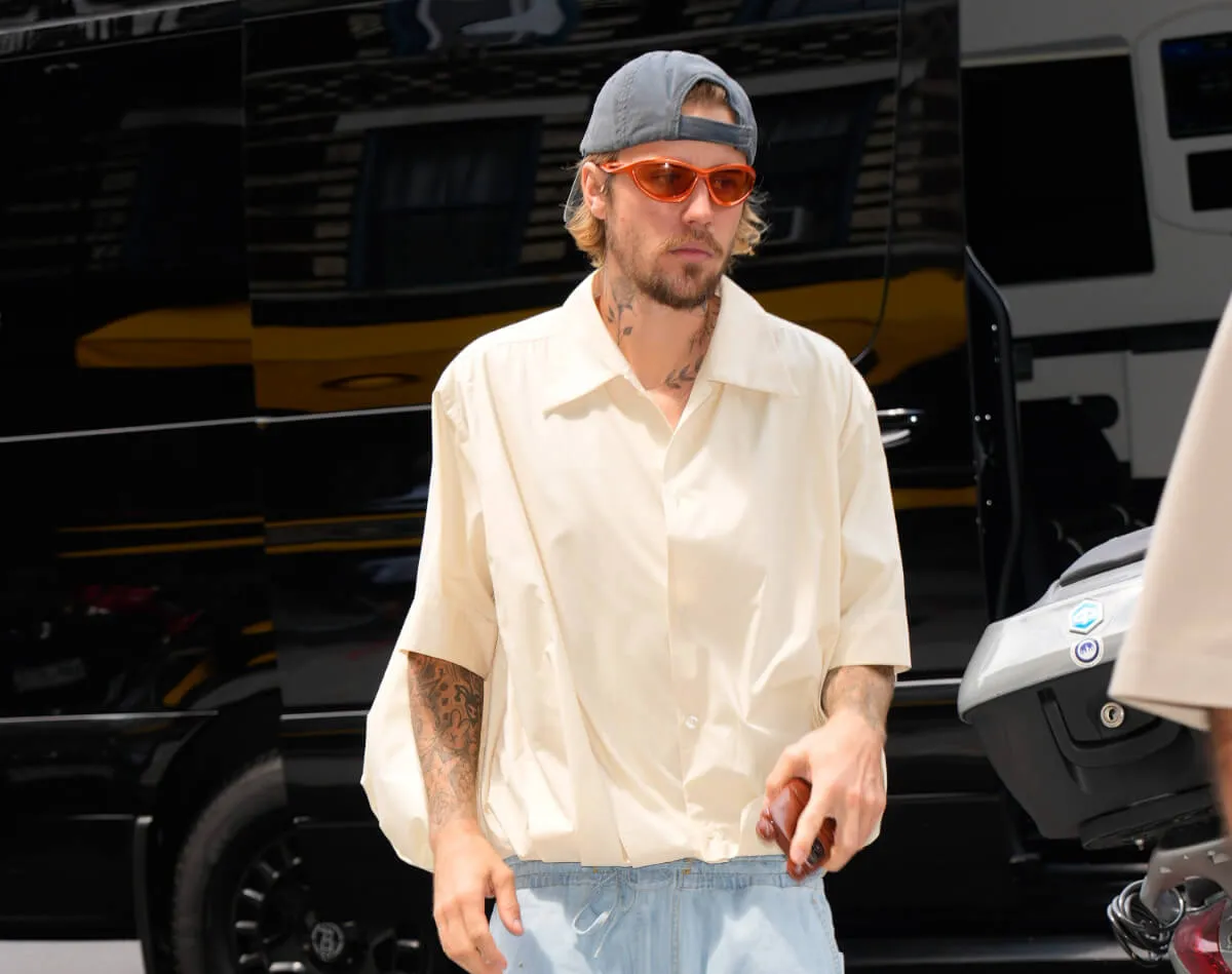 Justin Bieber wears orange sunglasses, a backwards baseball hat, and a white button down shirt.