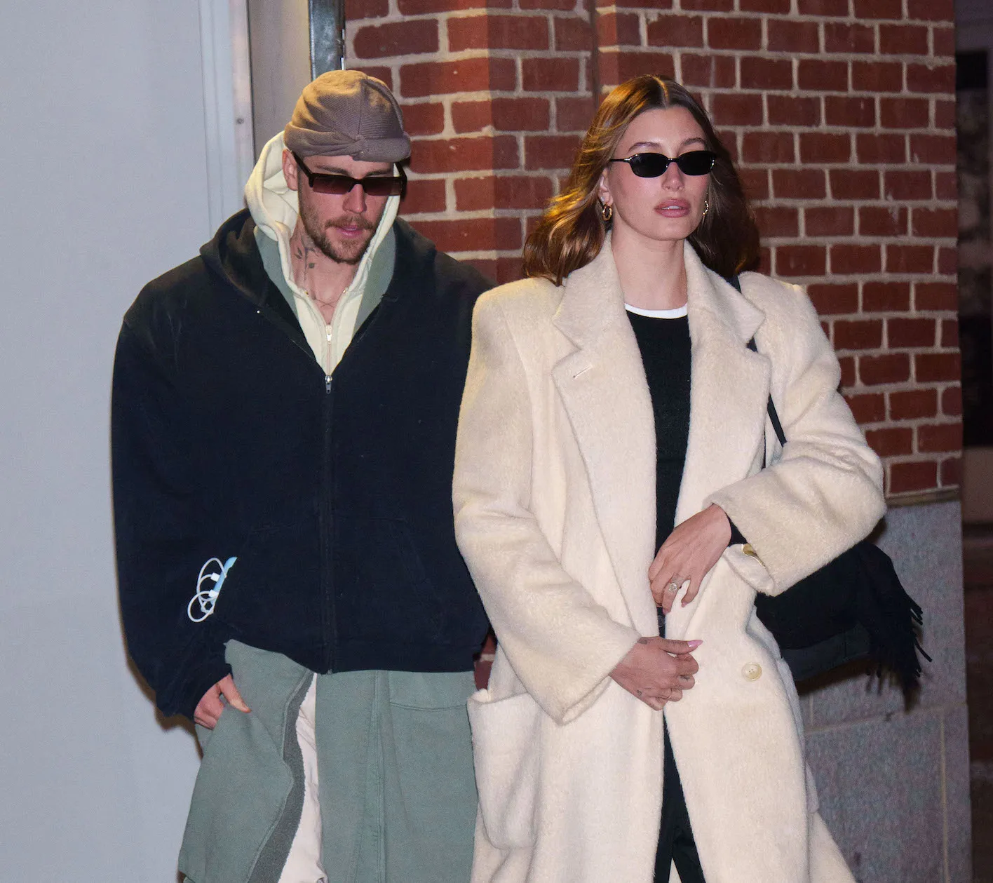 Justin Bieber and Hailey Bieber walking together in New York City in February 2025. Hailey is walking in front and wearing a cream coat and sunglasses. Justin is wearing a navy jacket over a long green jacket and cream pants.