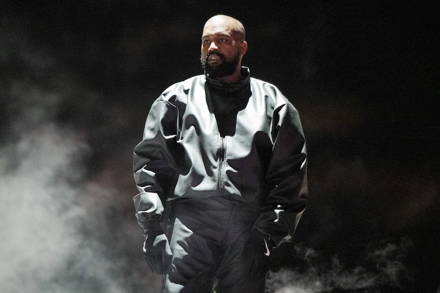 Kanye West wearing a black jacket against a black background on stage. He's surrounded by smoke.