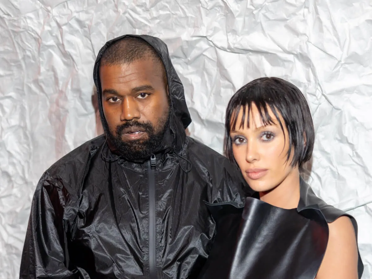 Kanye West wears a black jacket and stands with Bianca Censori, who wears a black tank top.