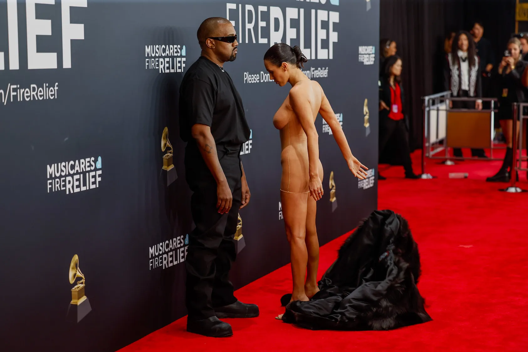 Kanye West and Bianca Censori on the red carpet at the Grammy Awards. West is wearing all black and facing the cameras. Censori is facing away from the cameras and dropping a black fur coat to reveal a nude look.