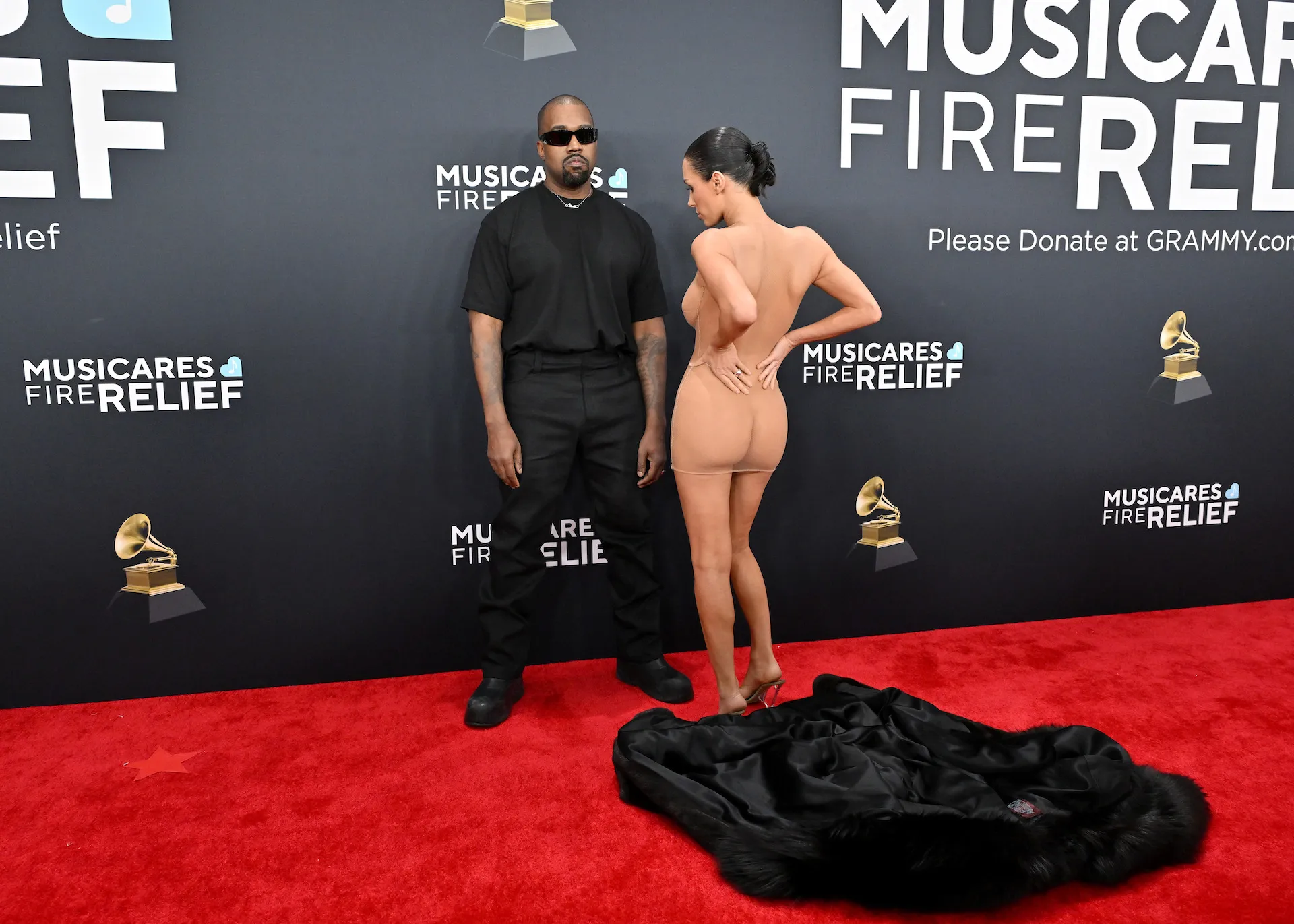 Kanye West and Bianca Censori on the red carpet at the Grammy Awards. West is dressed in black and Censori is nude with her back turned toward the cameras and her hands on her hips.