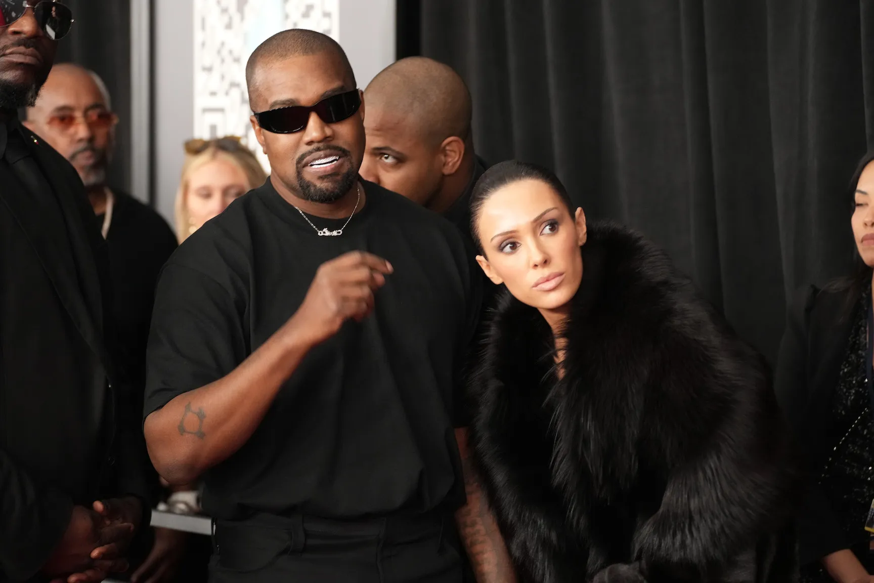 Kanye West smiling in all black with sunglasses on while next to Bianca Censori, who's in a black fur coat and leaning into him as she looks forward.