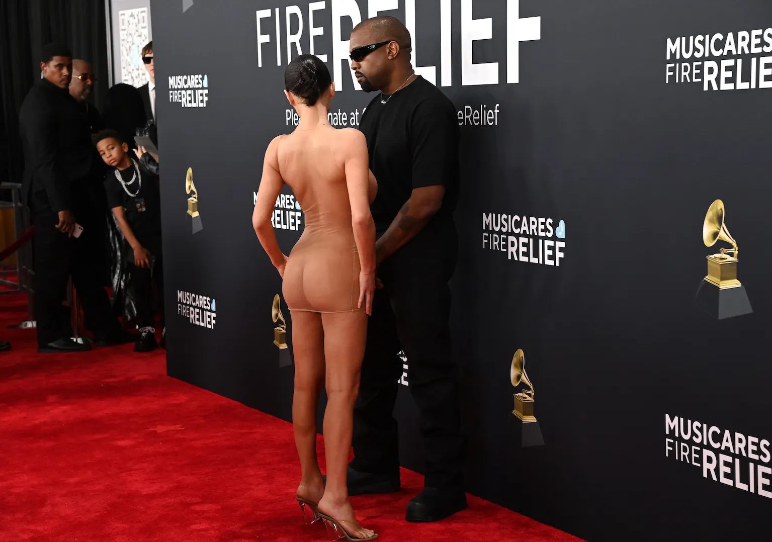 Kanye West standing beside Bianca Censori on the red carpet at the Grammy Awards. Censori is in a sheer oufit and facing away from the camera, and West is wearing all black.