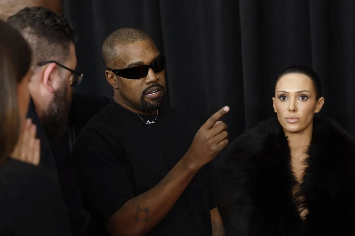 Kanye West speaking to someone as he and Bianca Censori attend the 67th GRAMMY Awards. He's pointing and wearing all black with black sunglasses.