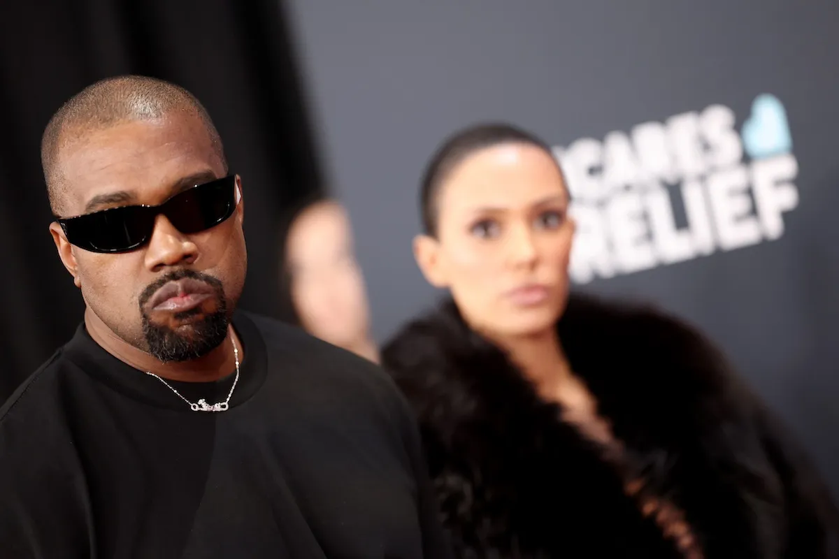 Kanye West wearing sunglasses and black T-shirt with out-of-focus Bianca Censori in the background