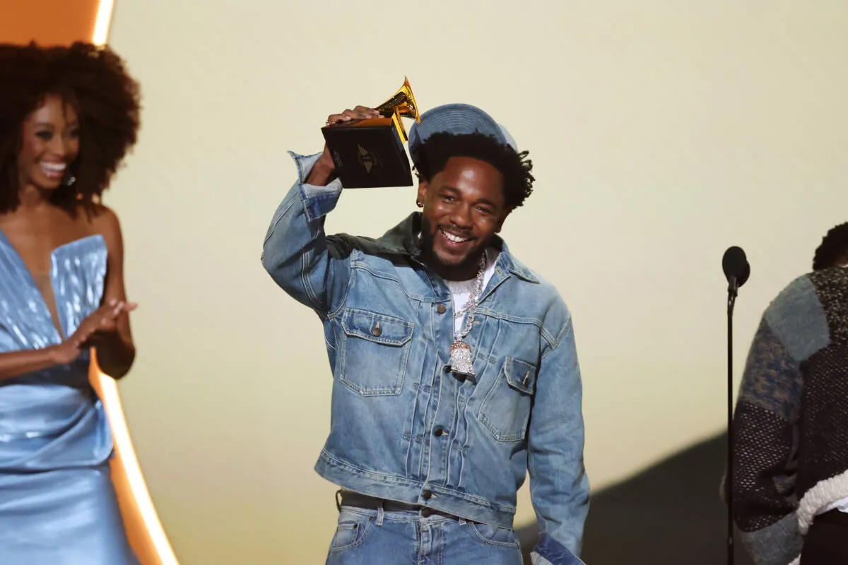 Kendrick Lamar wears a denim jacket and jeans and holds a Grammy up.