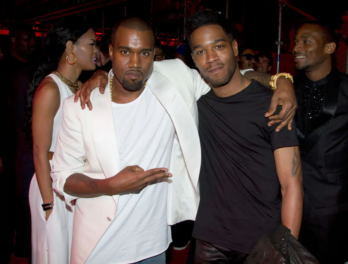 Kanye West and Kid Cudi stand with their arms around each other. West wears white and Cudi wears black.