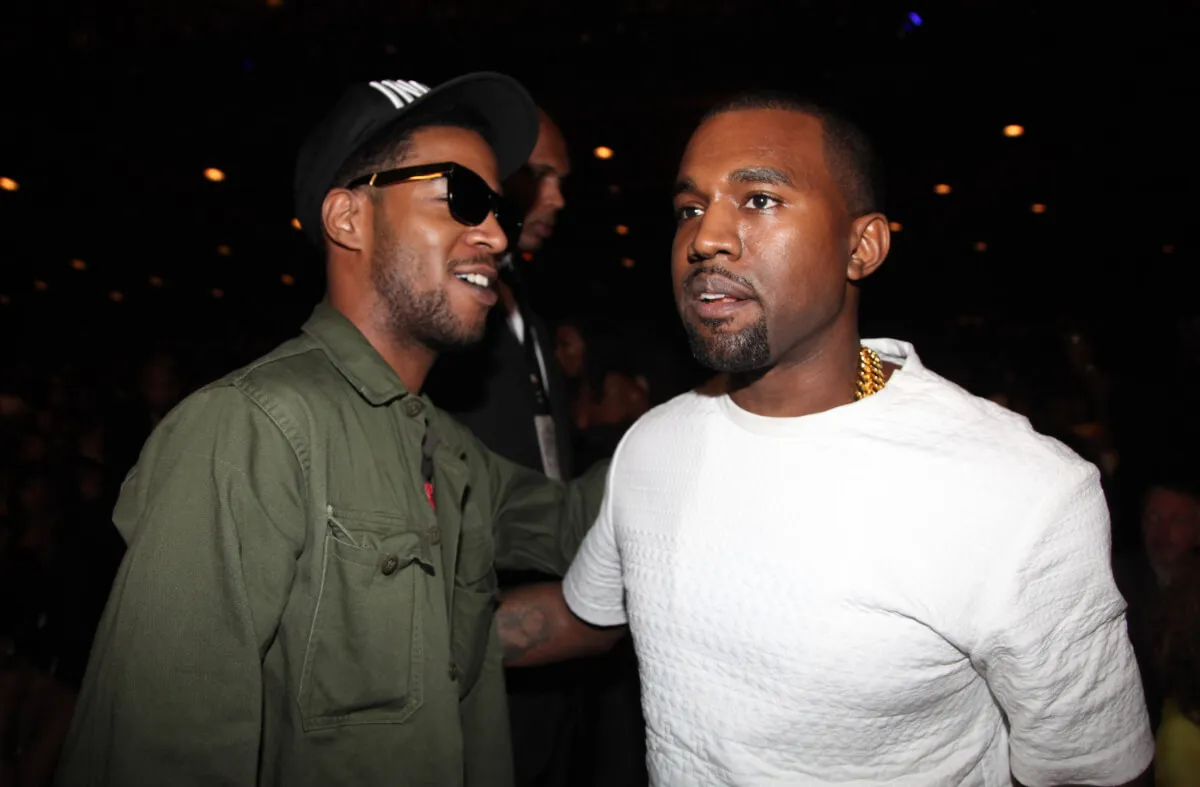 Kid Cudi wears a green jacket, sunglasses, and baseball hat. He stands with Kanye West, who wears a white shirt.