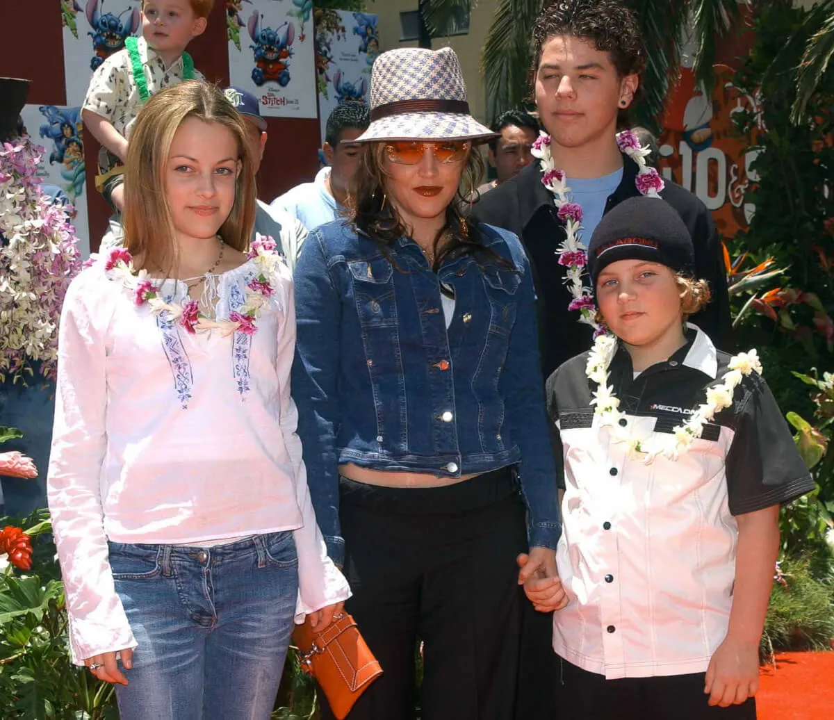 Riley Keough, Lisa Marie Presley, Navarone Garibaldi, and Benjamin Keough stand on a red carpet together. The children wear leis.