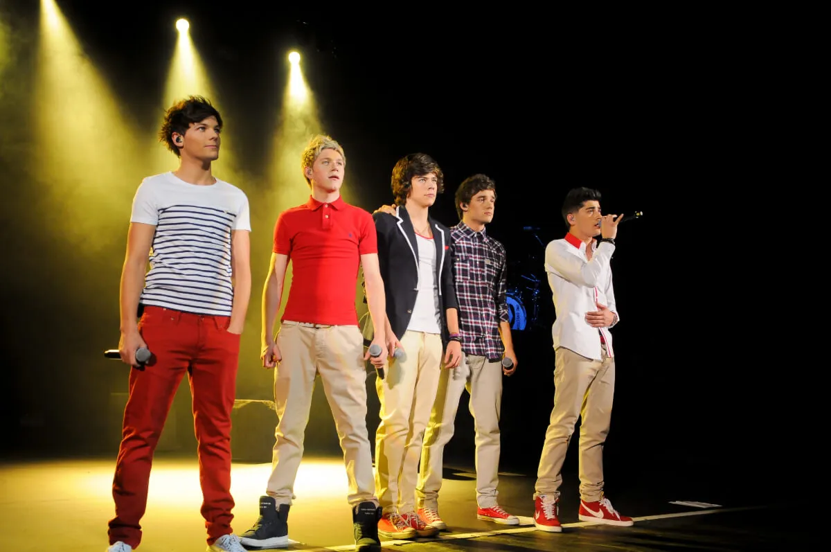 Louis Tomlinson, Niall Horan, Harry Styles, Liam Payne, and Zayn Malik stand in a line on a stage. They all hold microphones, and Malik sings into his.