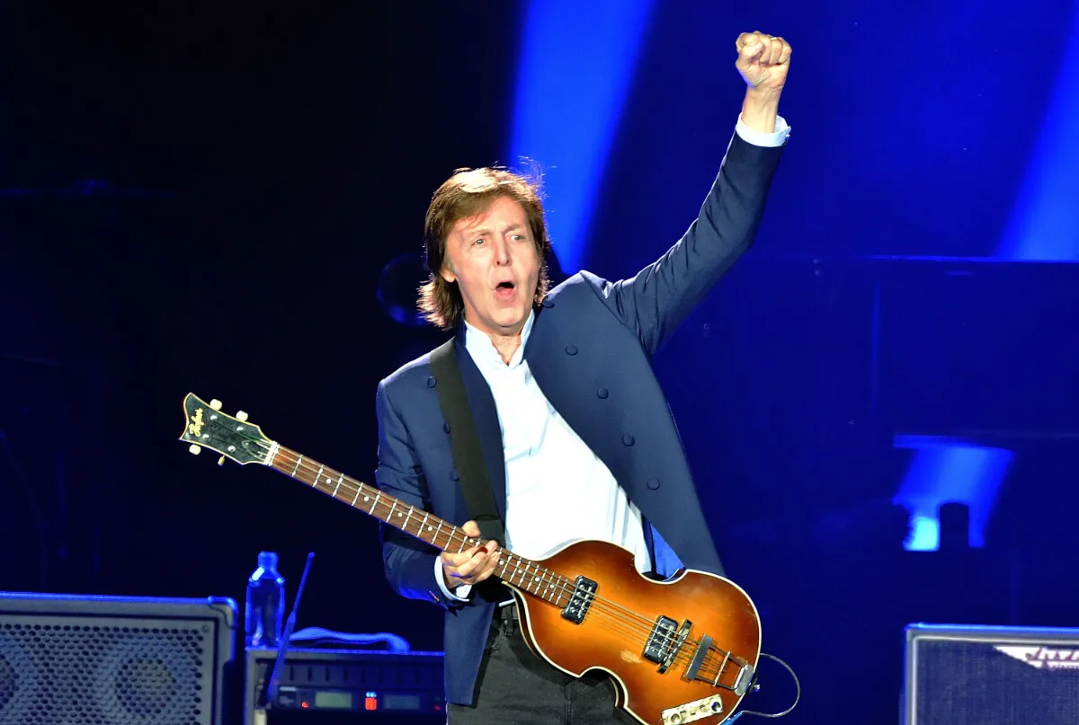 Paul McCartney holds a guitar and holds his fist up in the air.