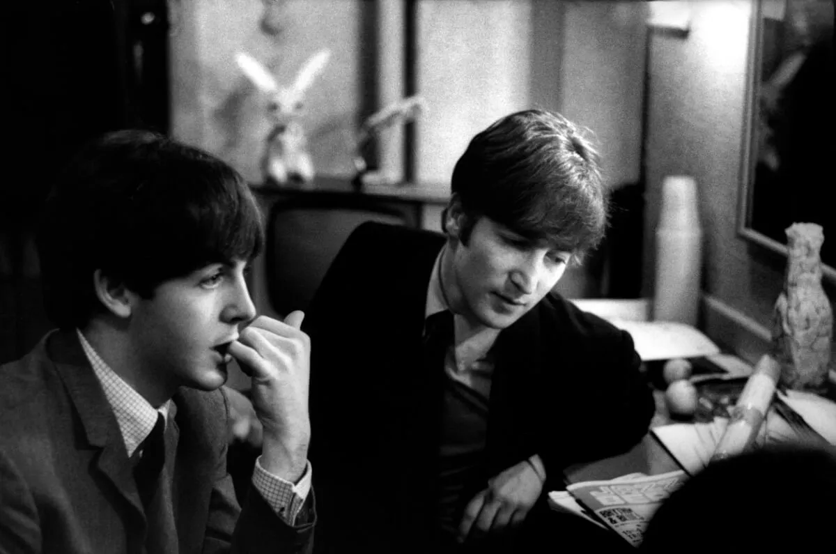 A black and white picture of Paul McCartney and John Lennon sitting together wearing suits. McCartney sits in profile and Lennon looks at something on a desk.