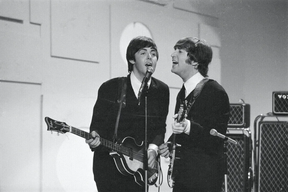 A black and white picture of Paul McCartney and John Lennon playing guitars and singing into the same microphone. They wear suits.