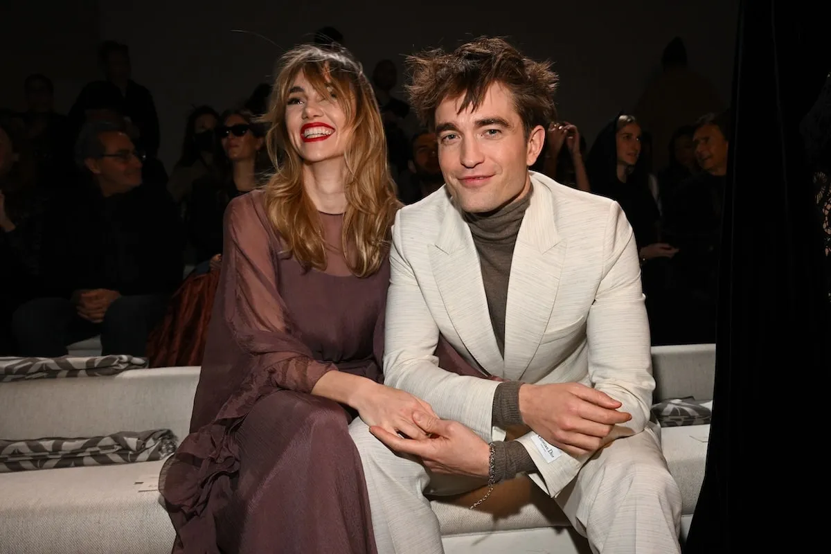 Wearing a purple dress and white suit respectively, Suki Waterhouse and Robert Pattinson smile together in the front row of a 2023 Dior fashion show