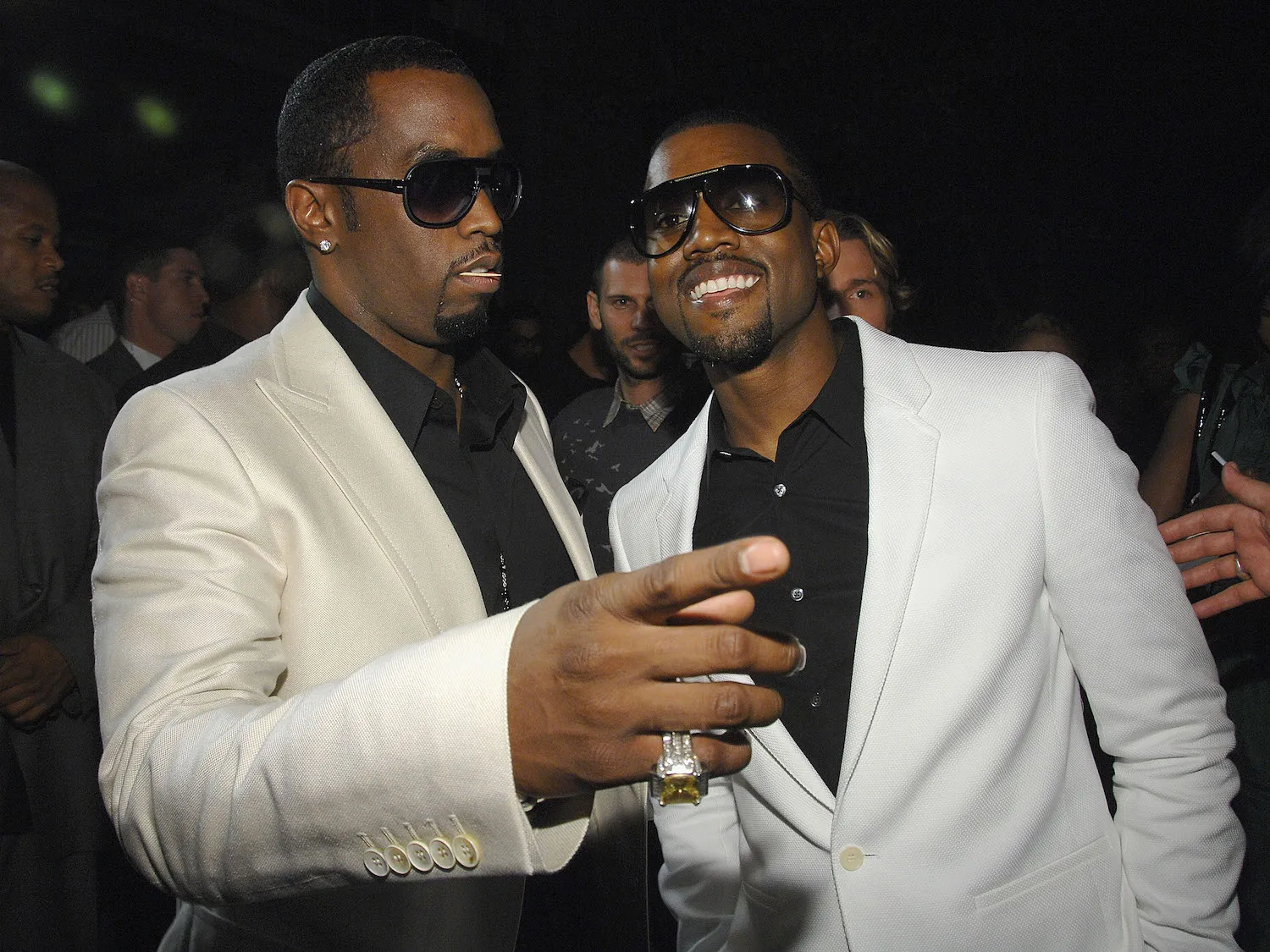 Sean 'Diddy' Combs in a cream-colored suit pointing with Kanye West in a white suit smiling next to him 