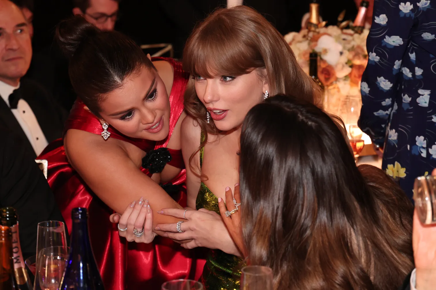 Selena Gomez leaning over Taylor Swift's shoulder and talking to someone next to Swift at the Golden Globes.