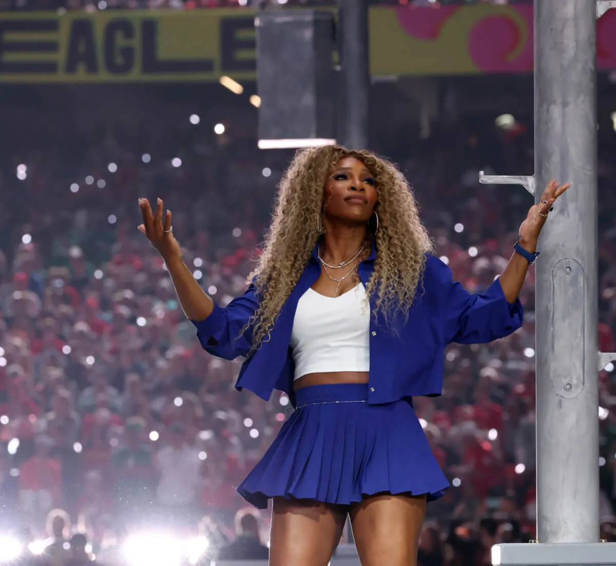 Serena Williams wears a blue tennis skirt and jacket. She stands with her arms up in a shrug at the Super Bowl.