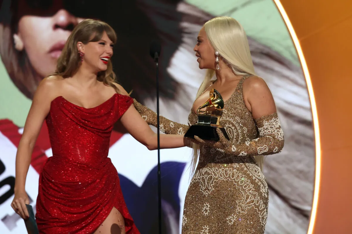 Taylor Swift and Beyonce stand together on stage. Beyonce holds a Grammy. Swift wears a red dress and Beyonce wears a gold dress and gloves.