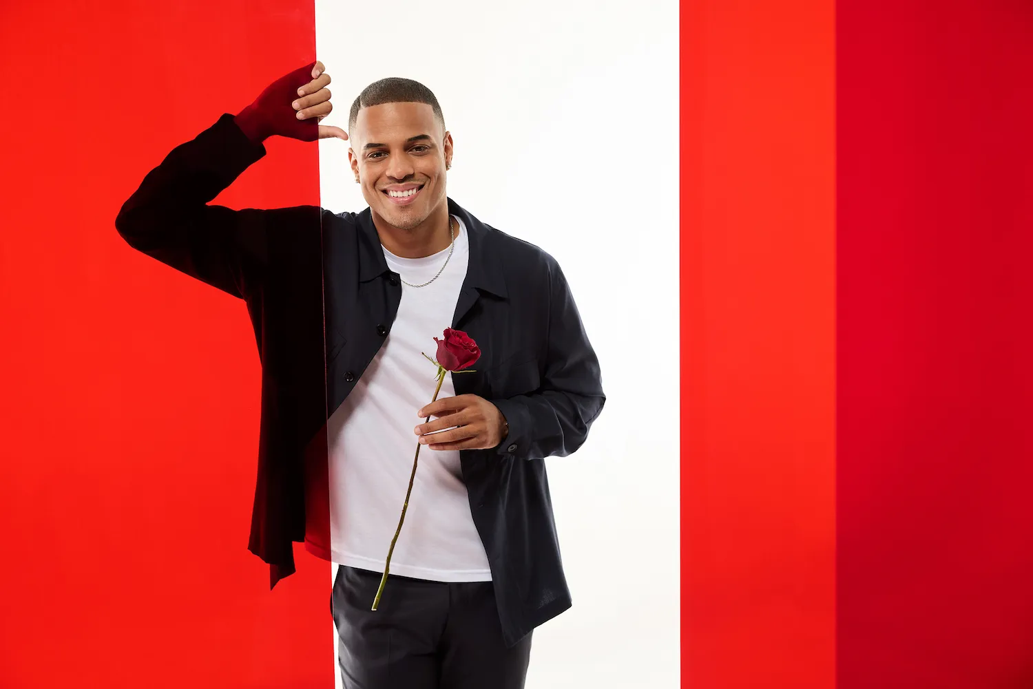 'The Bachelor' Season 29 lead Grant Ellis holding a rose and leaning against a red wall. He's against a red and white background.