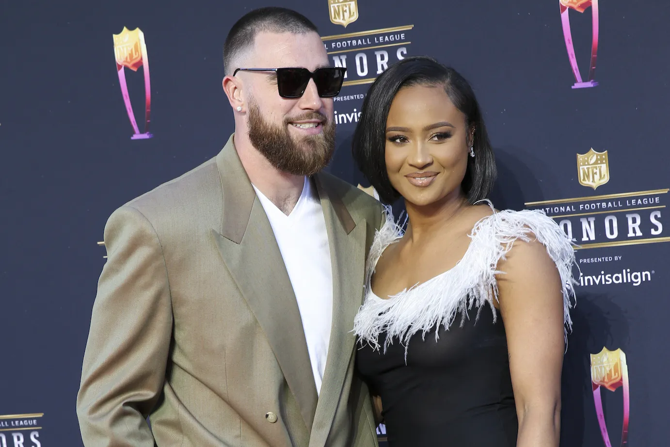 Travis Kelce wearing sunglasses and smiling next to Kayla Nicole, who's wearing a fur-lined dress and smiling